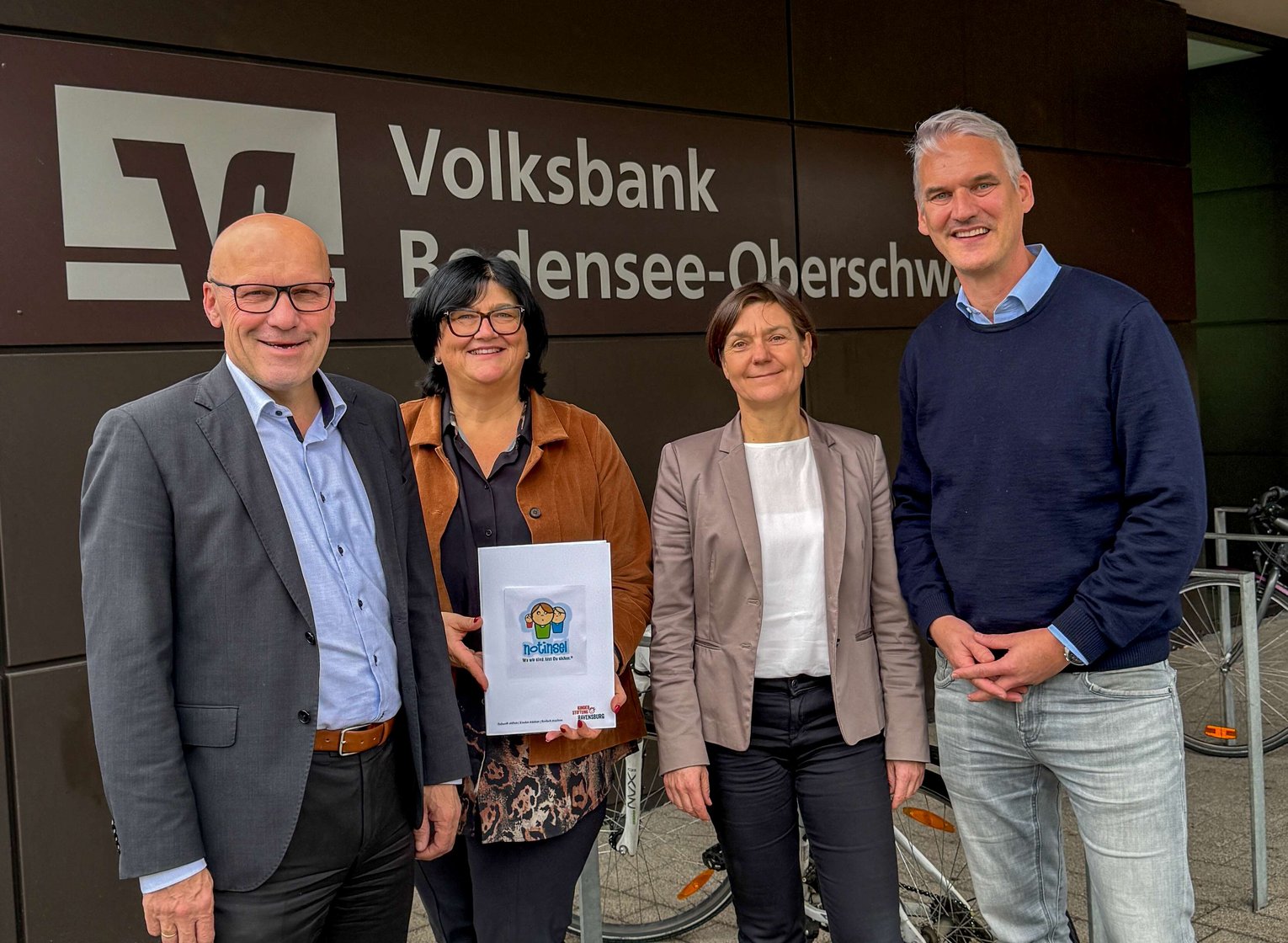 Vier Personen vor dem Eingang der Hauptgeschäftsstelle Ravensburg mit dem Logo von Notsinsel.